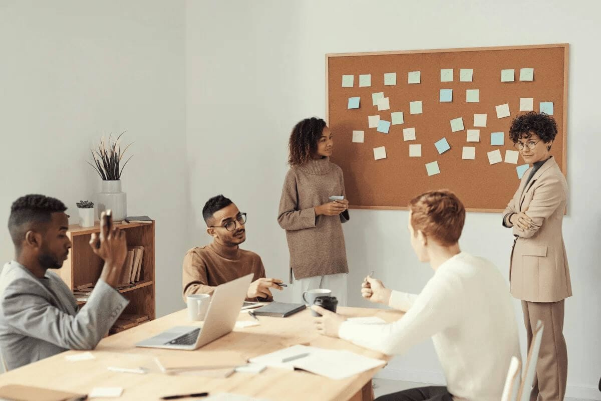 Team brainstorming session with post-it notes on a corkboard, discussing ideas around a table.