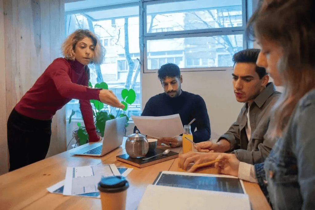 Team discussing documents around a table in a casual meeting room with a window view.