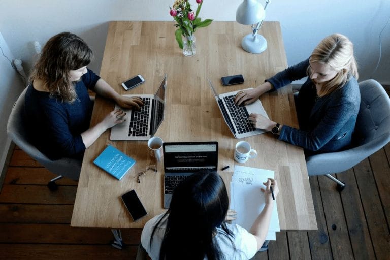 Three women collaborating on laptops with coffee cups and notebooks.