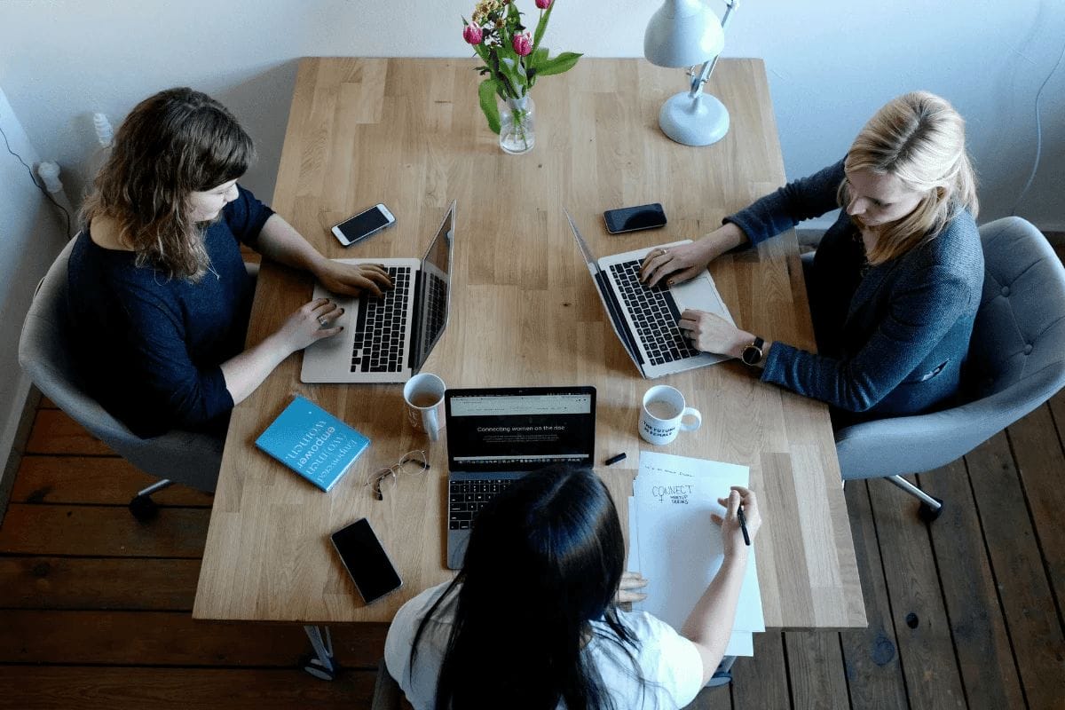 Three women collaborating on laptops with coffee cups and notebooks.