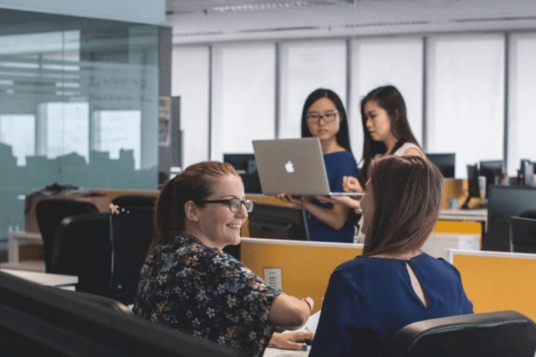 Group of employees discussing work while using laptops in an open-plan office.