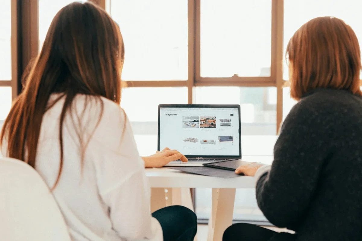 Two women working on a laptop with a website displaying furniture.