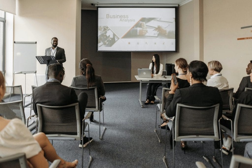Business presentation with a speaker at a podium and an audience seated in a conference room.