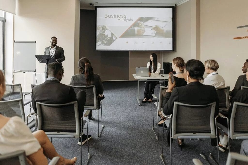 Professional business meeting with attendees listening to a speaker and a business analysis slide.