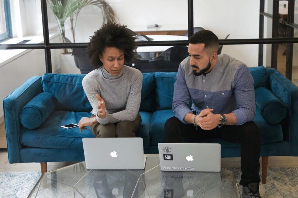 Two individuals seated on a couch, each using a laptop, engaged in a focused discussion or work session.
