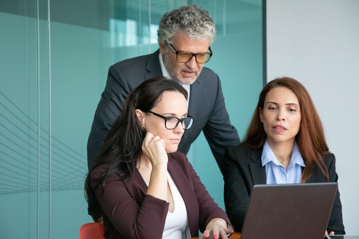 Three business professionals engaged in a discussion around a laptop in a modern meeting room.