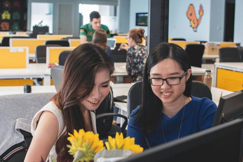 Two women collaborating on computers in a modern office setting, focused on their tasks and engaged in work.
