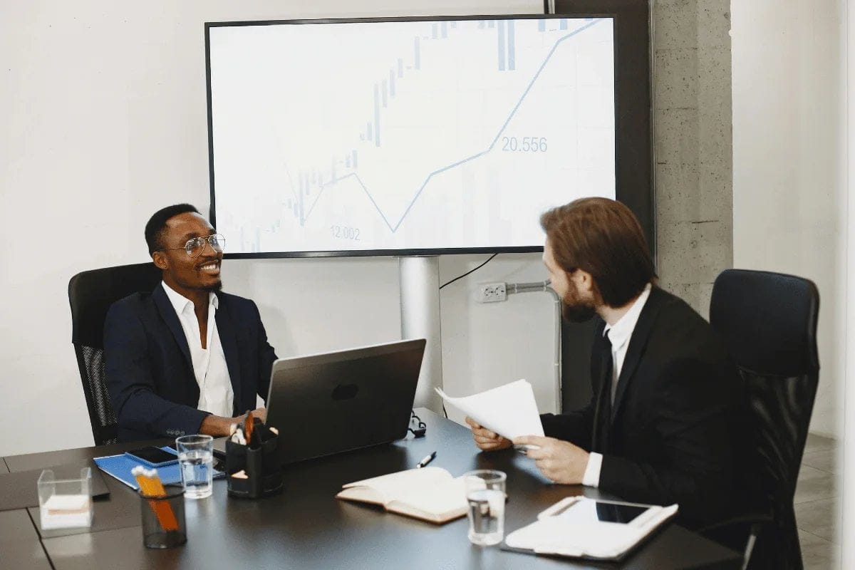 Two men in business suits seated at a table, engaged with a laptop in a professional setting.
