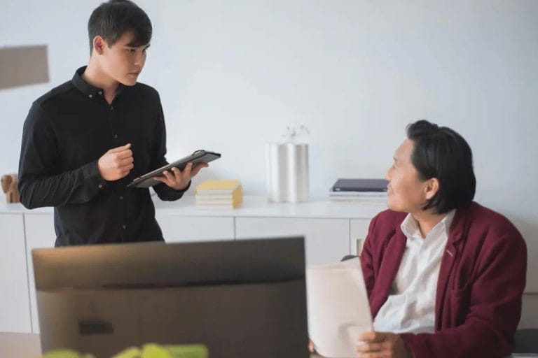 A business professional discusses a document with a colleague, holding a tablet in an office setting.