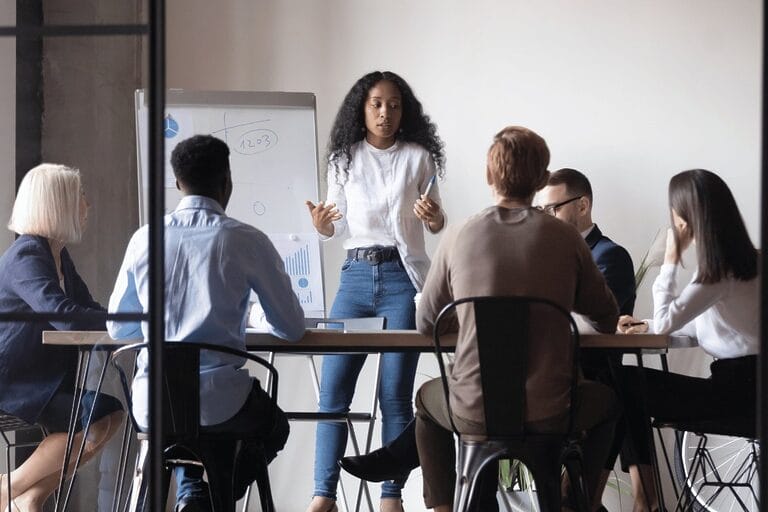 A diverse group of people in a professional setting, sitting around a table with notebooks and laptops, engaged in a discussion or collaborative activity.