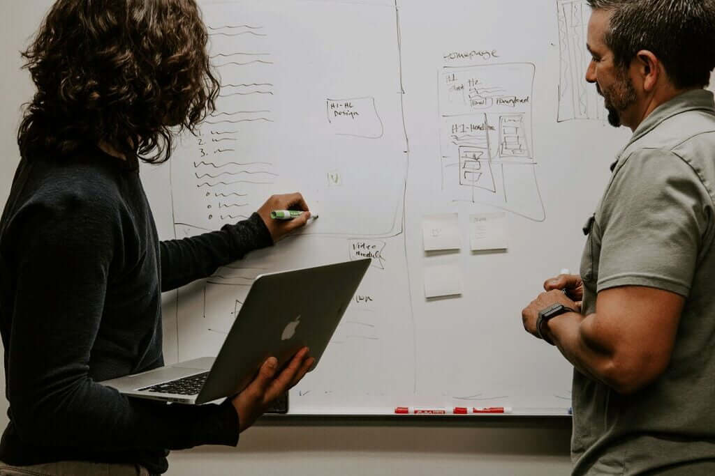 Two professionals standing in front of a whiteboard, engaged in discussion and pointing at various notes and diagrams.