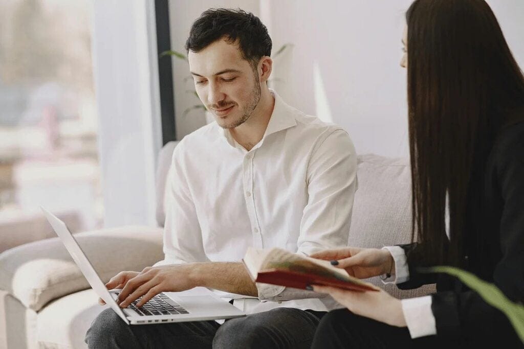 Two professionals with a laptop and a book sitting in a bright office setting.