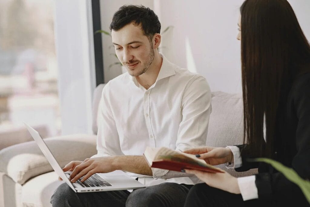 Two professionals with a laptop and a book sitting in a bright office setting.