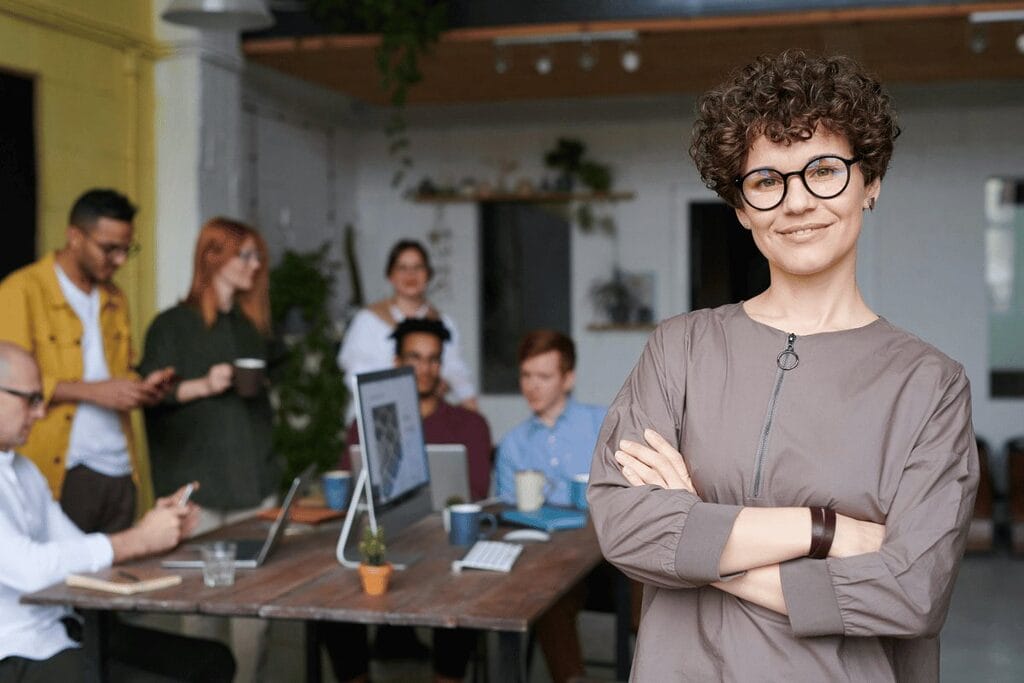 Person with crossed arms in front of a group meeting in a casual office setting.