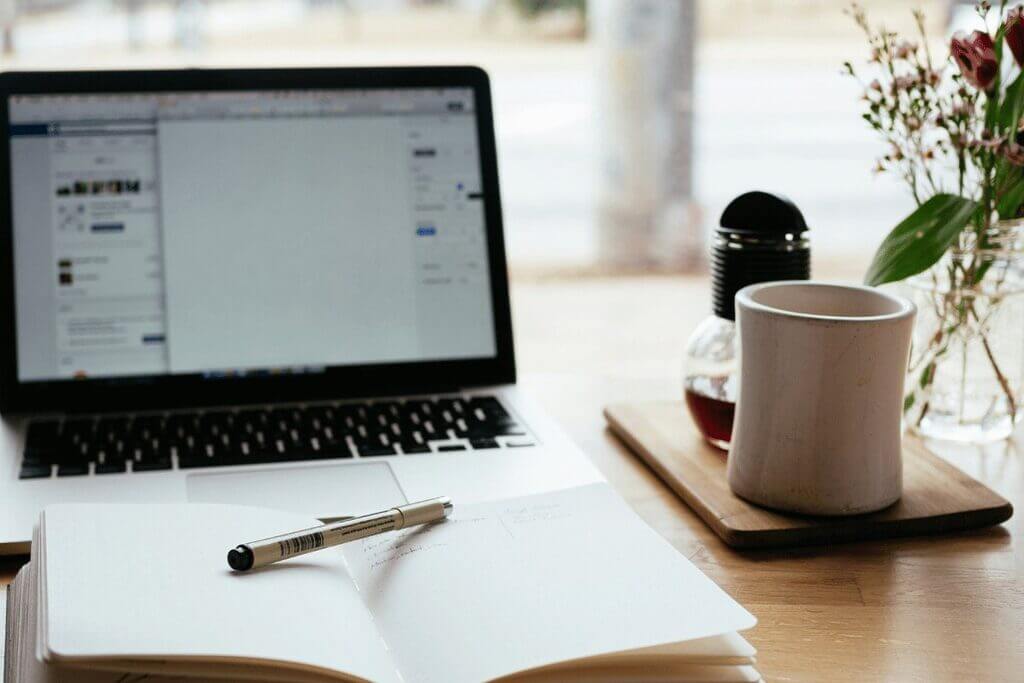 A laptop, notebook, and cup of coffee arranged neatly on a rustic wooden table, creating a cozy workspace atmosphere.
