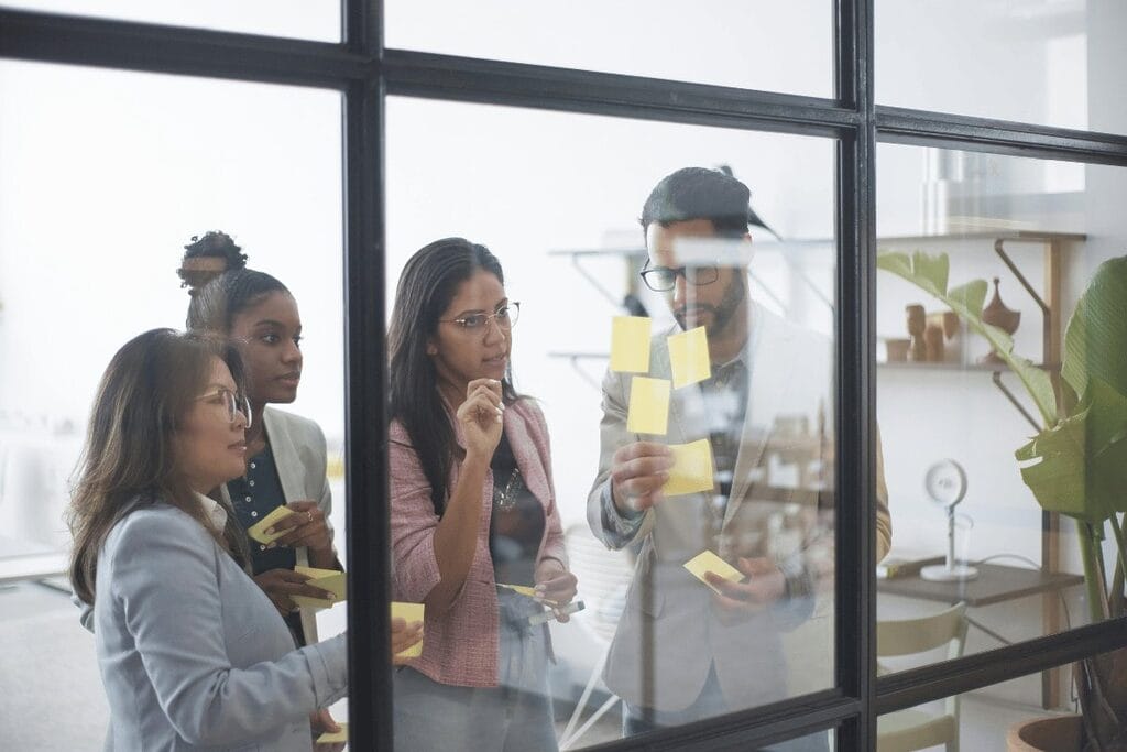 Three individuals in an office setting collaborating over sticky notes on a glass window.