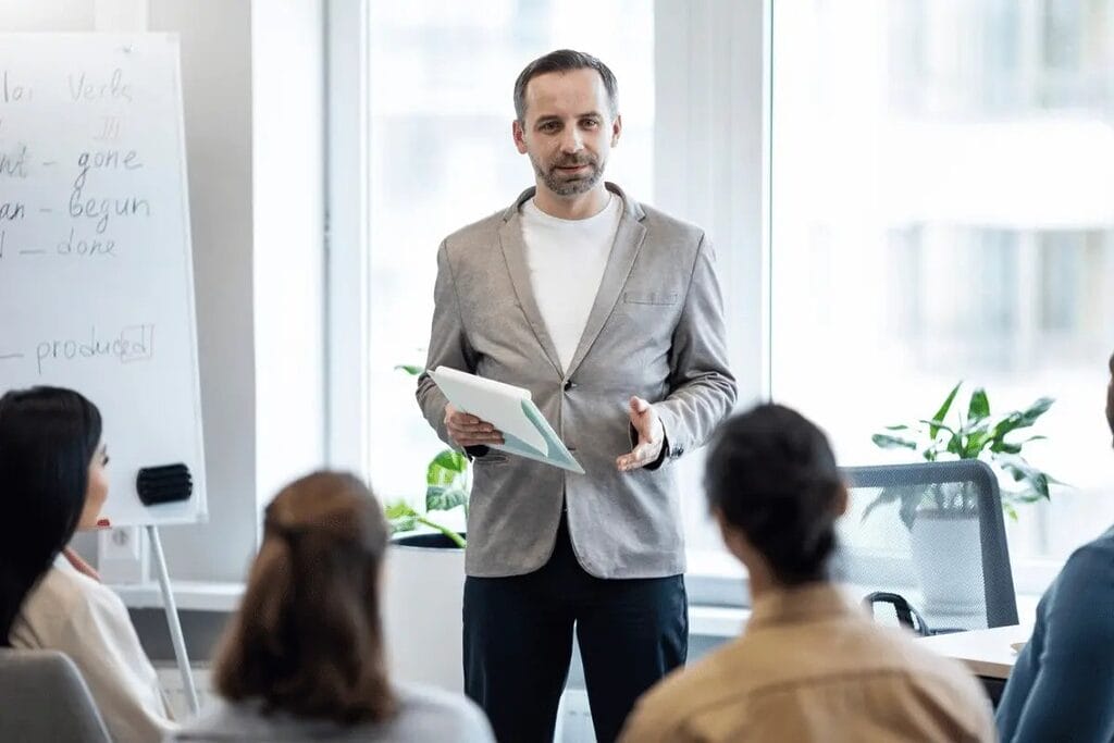 Man presenting to a group of people in an office with a whiteboard in the background.