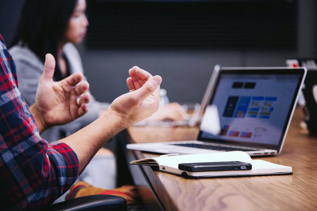 Person gesturing during a meeting with a laptop and smartphone on the table.