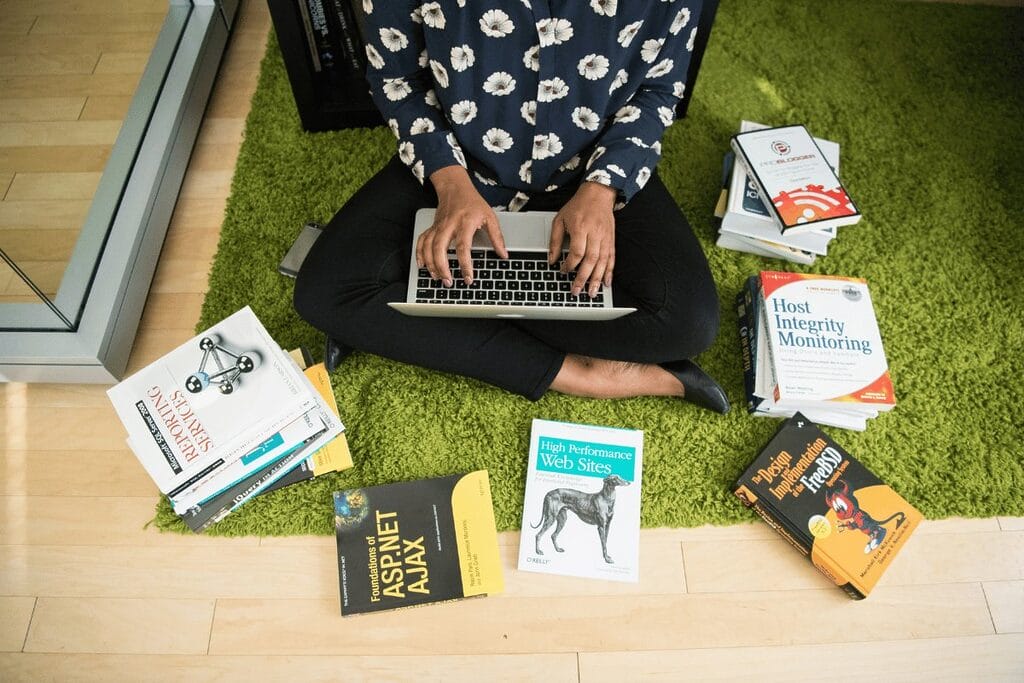 Person sitting on floor with laptop surrounded by scattered books.