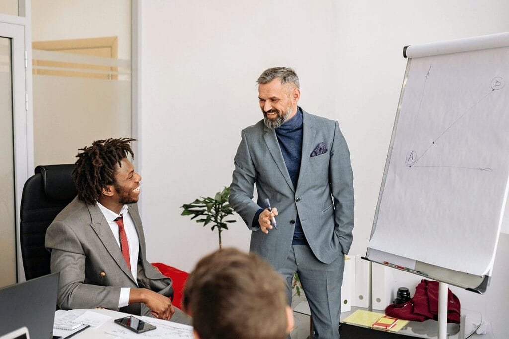 Two professionals in a meeting room with one person standing by a whiteboard.