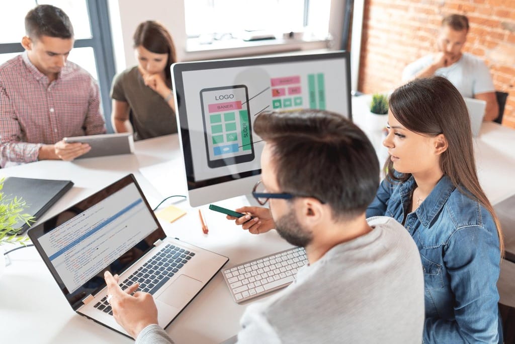 Professionals in a meeting with laptops and a monitor displaying a mobile app design.