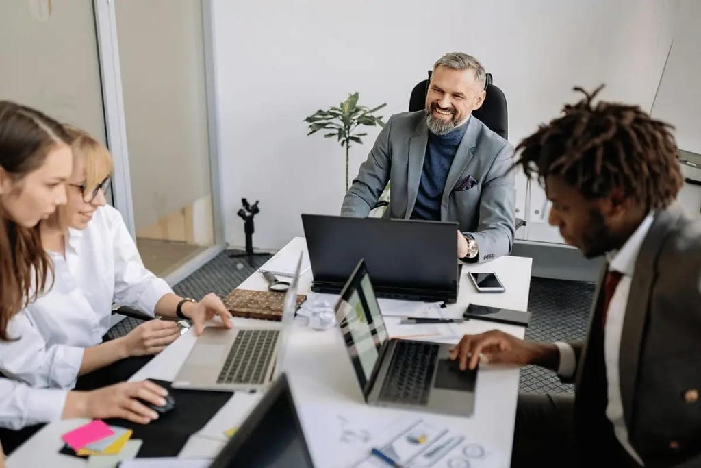 Group of colleagues in a business meeting, focused on their laptops, led by a smiling man.