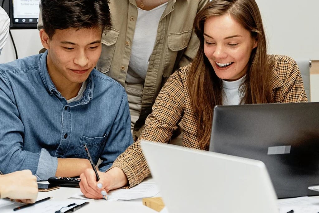 Two colleagues collaborating on a project, smiling and using a laptop at a shared desk.