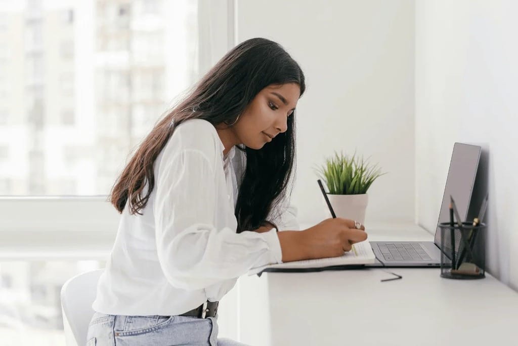 Professional woman writing in a notebook in front of a laptop.