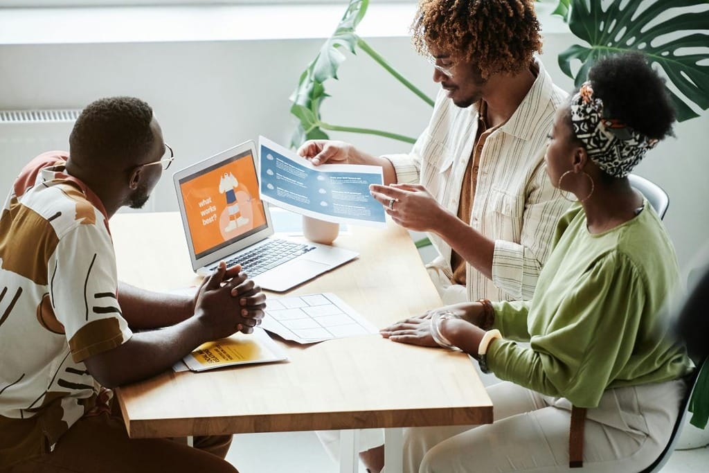 Three people at a table with a laptop and documents, discussing work.