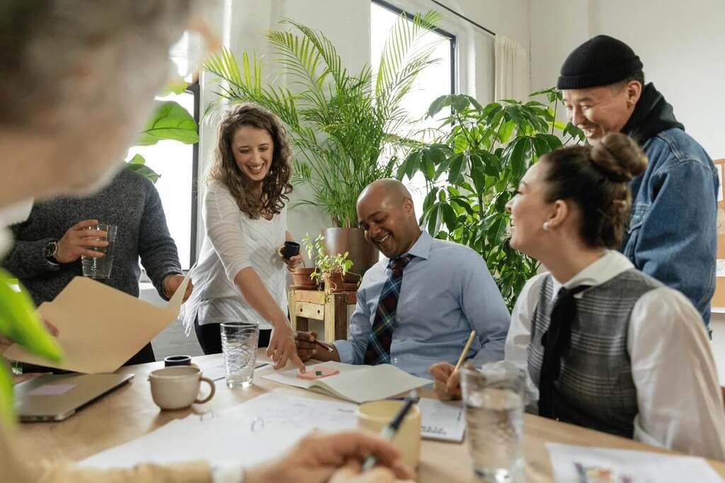 A diverse team laughing and collaborating around a table with plants in the background.