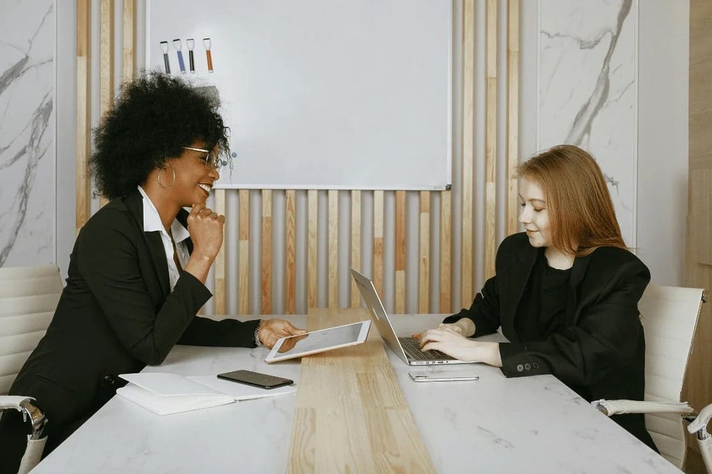 Woman holding a tablet while talking to a coworker at a desk.