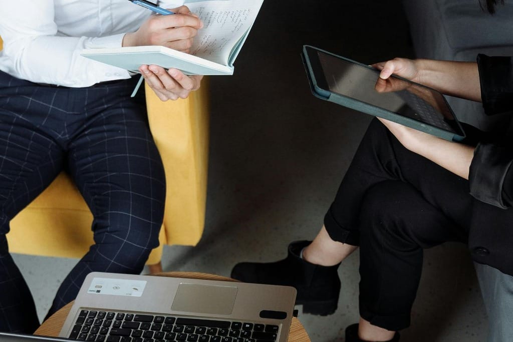 Two people in a meeting with a notebook, tablet, and laptop on a table.