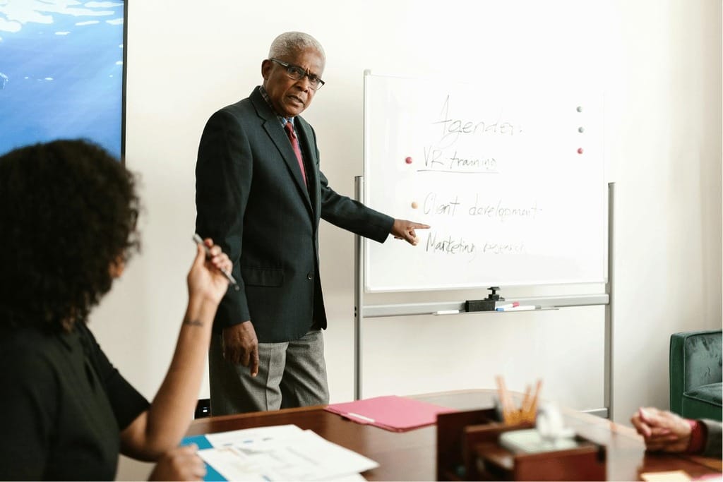 Senior executive pointing at a whiteboard with a meeting agenda in a business discussion.