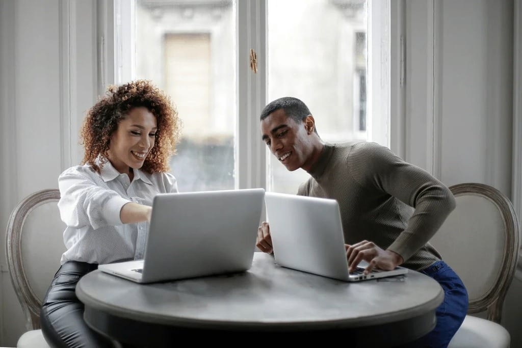Office setting with two people engaged in a friendly discussion while working on laptops.