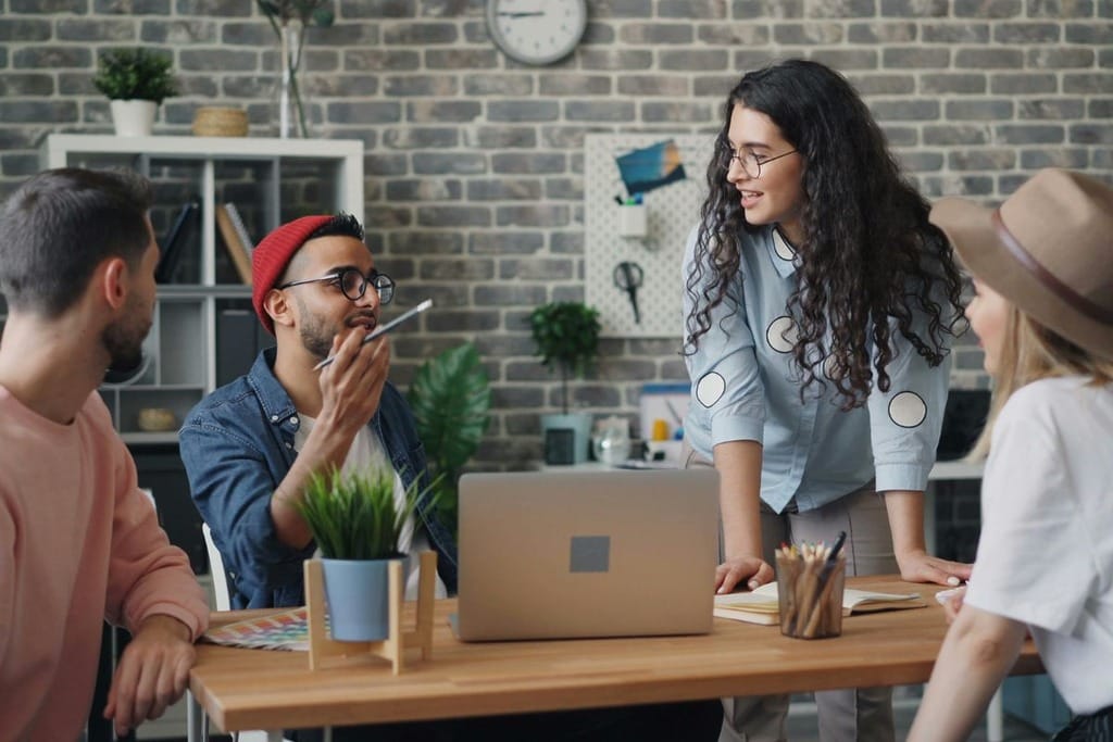 Four colleagues in a casual meeting around a wooden table with a laptop and stationery.