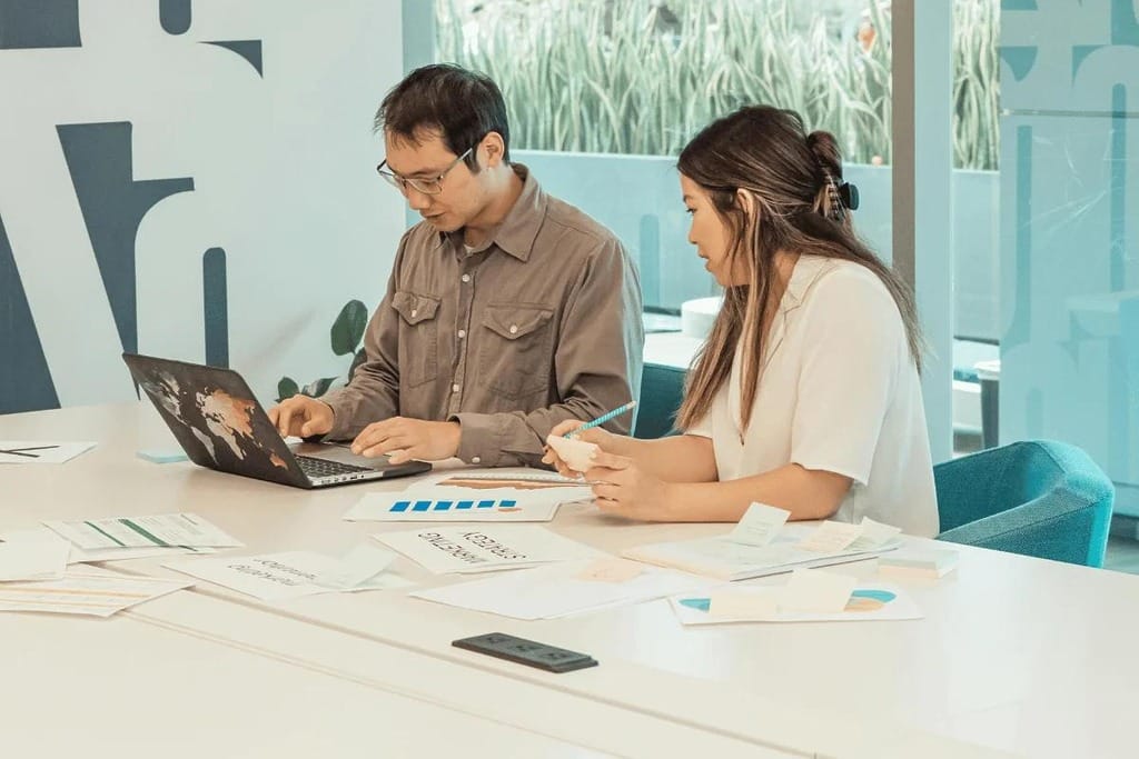 Coworkers discussing work over a laptop and papers in a bright office.