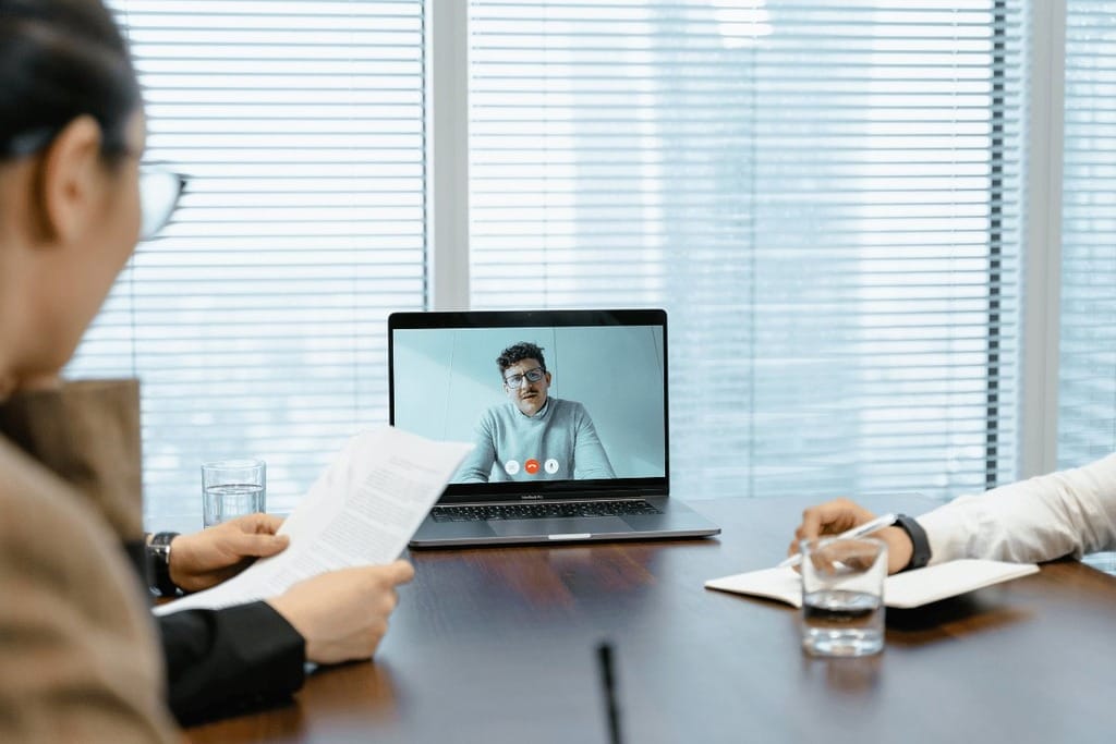 Two individuals in a video call meeting, one on a laptop screen, in a modern office setting.