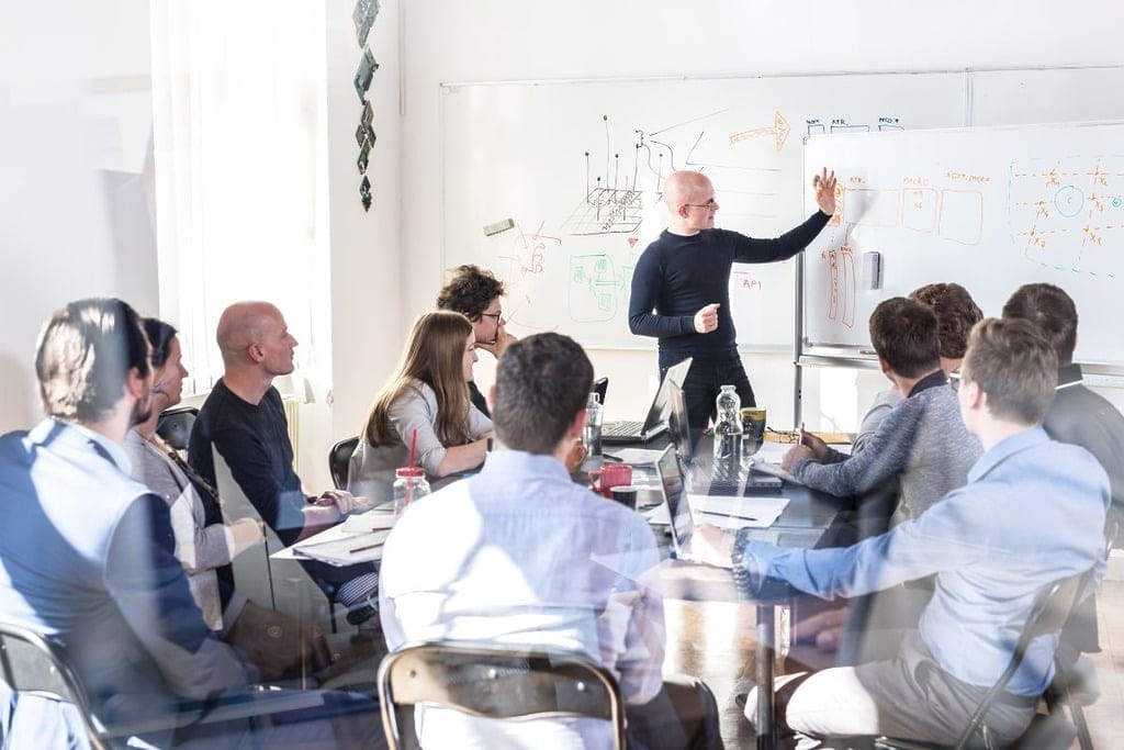 Group of professionals in a meeting with one person presenting at a whiteboard.