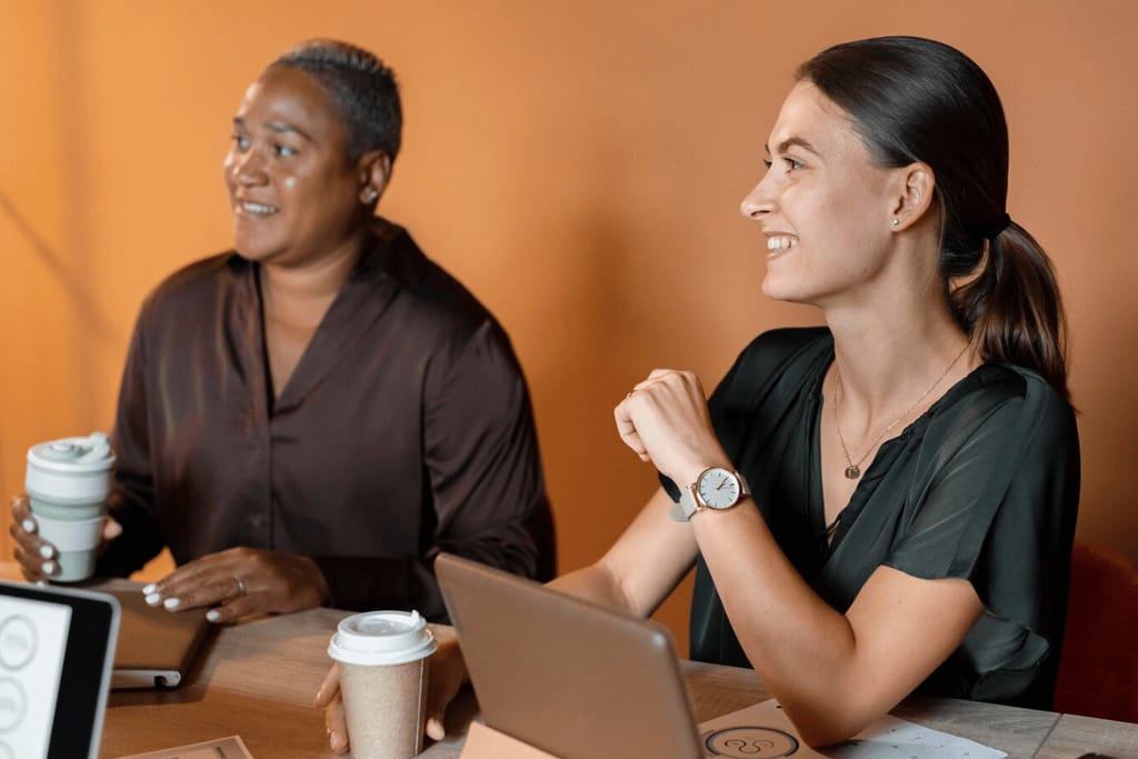 Two businesswomen smiling during a meeting with laptops and coffee.