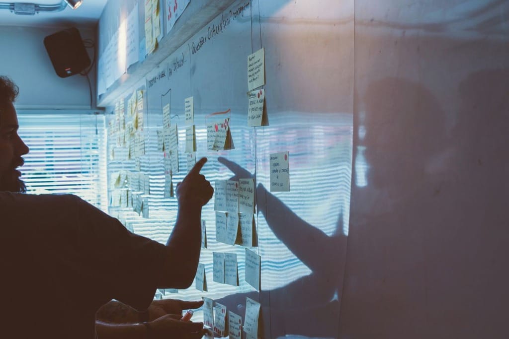 A man points to a whiteboard covered in colorful sticky notes during a presentation or brainstorming session.