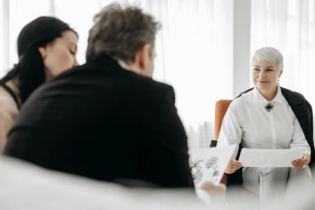 A group discussion takes place in a bright, modern room, with two individuals listening attentively to a seated person holding documents.
