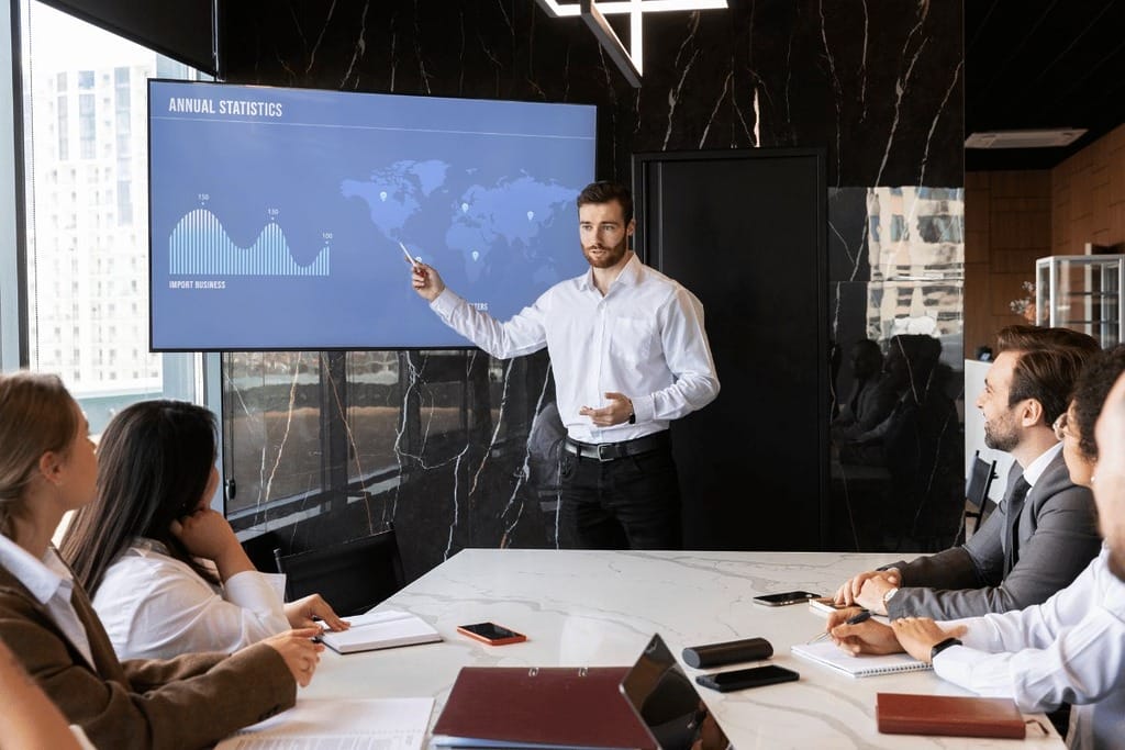 A presenter points to a large screen displaying annual statistics during a business meeting, while audience members attentively listen.