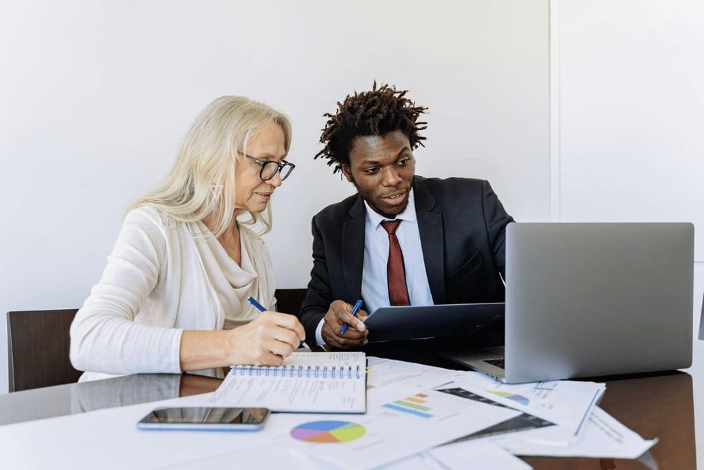 Two individuals dressed in business attire working together at a table with a laptop open between them.