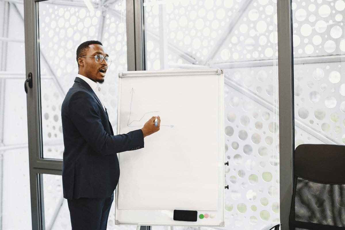 A man in a suit stands in front of a whiteboard, illustrating a chart in a modern office with a patterned wall and a chair visible.