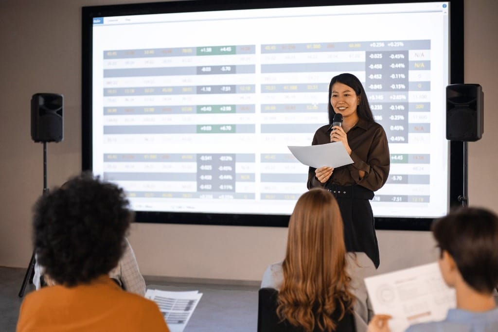 A speaker presents data from a large screen to an audience, who are actively listening and taking notes.